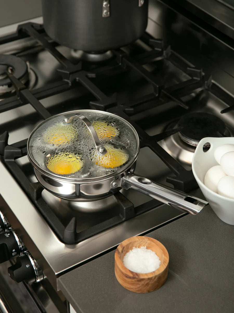 Eggs being cooked in a pan on a gas stove with a bowl of salt nearby.
