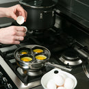 Person cracking eggs into a four-sectioned egg poacher on a stovetop.