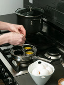 Person cracking eggs into a metal egg poacher on a stove with a bowl of eggs nearby.