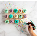 Cupcakes arranged to spell 'Happy Birthday' on a marble surface with colorful sprinkles and a person using an icing pen to add an exclamation mark to the final cupcake.