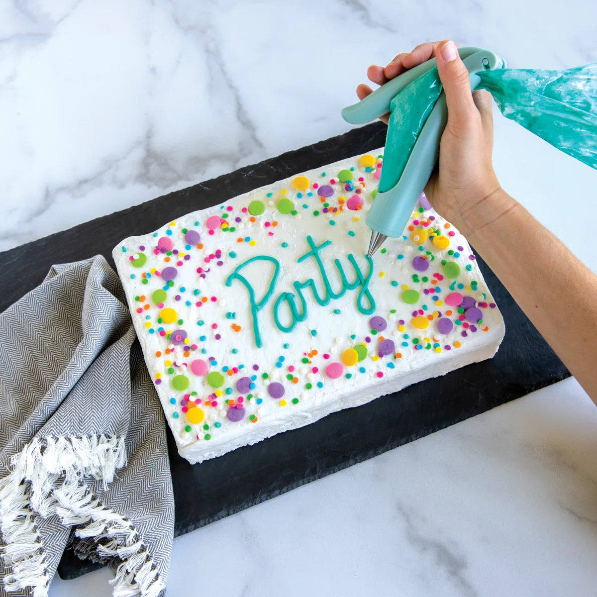 Person decorating a cake with colorful sprinkles and the word 'Party' on a marble surface.