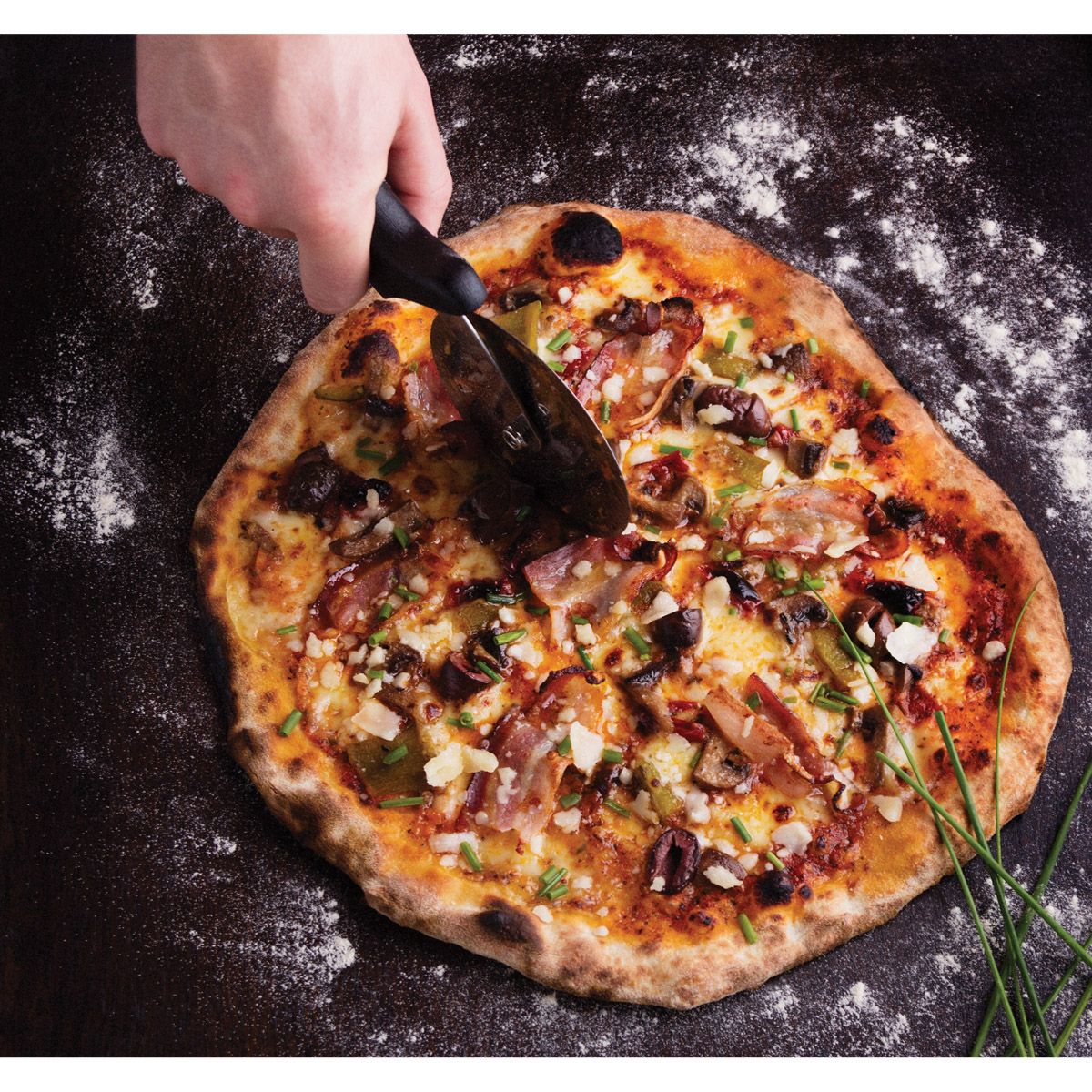 A person using a pizza cutter to slice a pizza on a dark background with flour sprinkled around it.