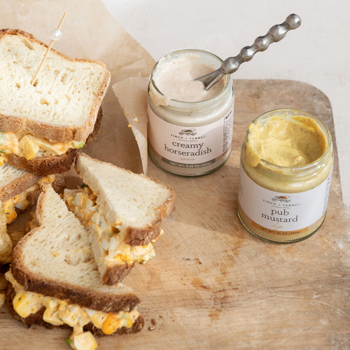 Sandwiches with mustard on a wooden board next to jars of creamy horseradish and pub mustard.