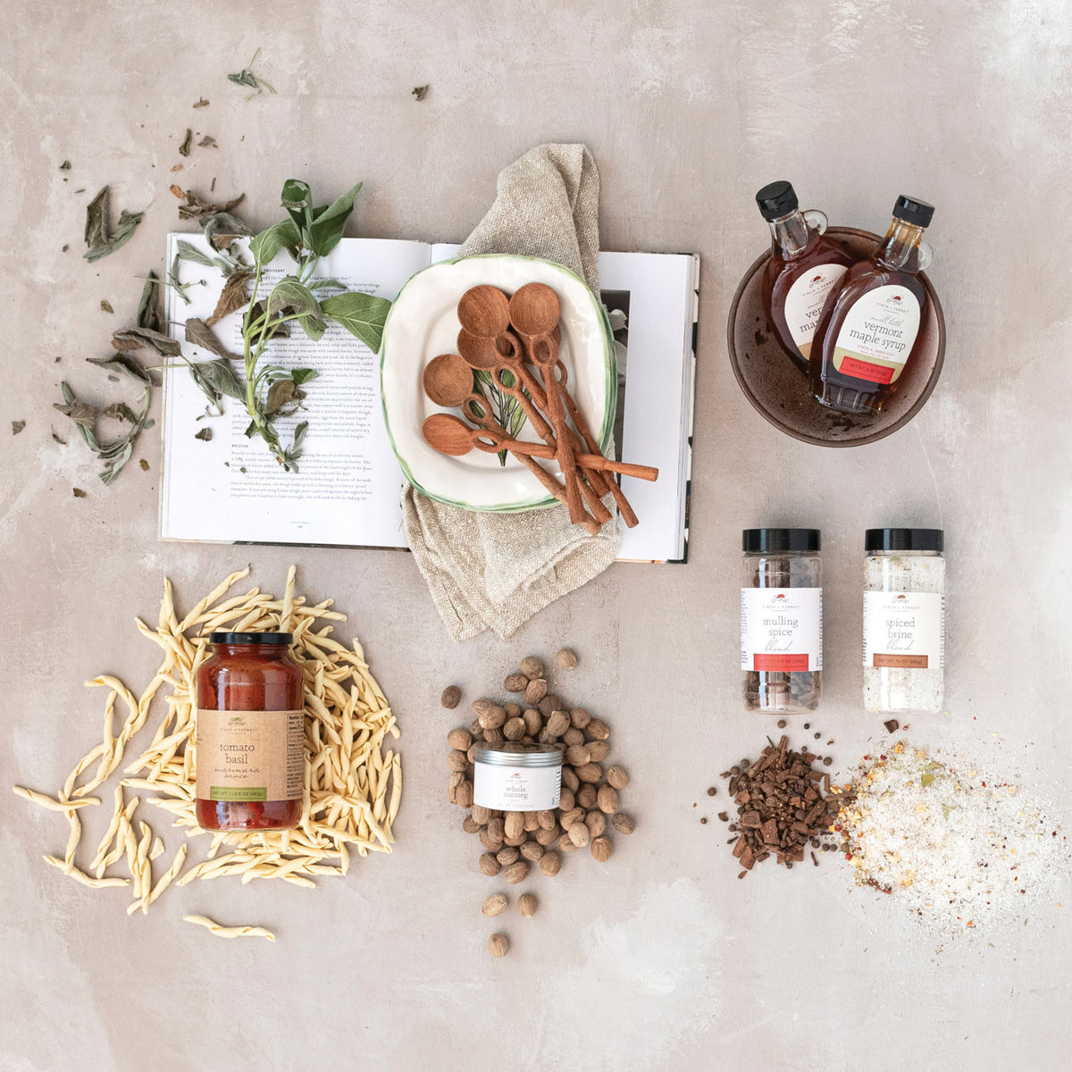 An overhead flat-lay of a gourmet food or spice gift set on a textured light gray surface. The set includes a jar of honey, two bottles of syrup, two spice jars, a tin of nut butter, loose spices, cinnamon sticks, dried herbs, and an open book with a cloth napkin.