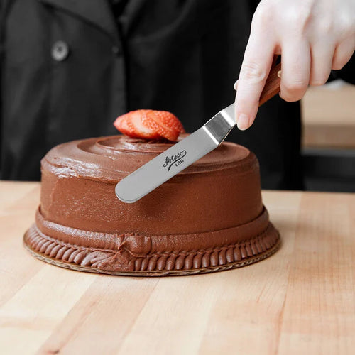Person using a cake knife to spread chocolate frosting on a small chocolate cake with a strawberry on top.