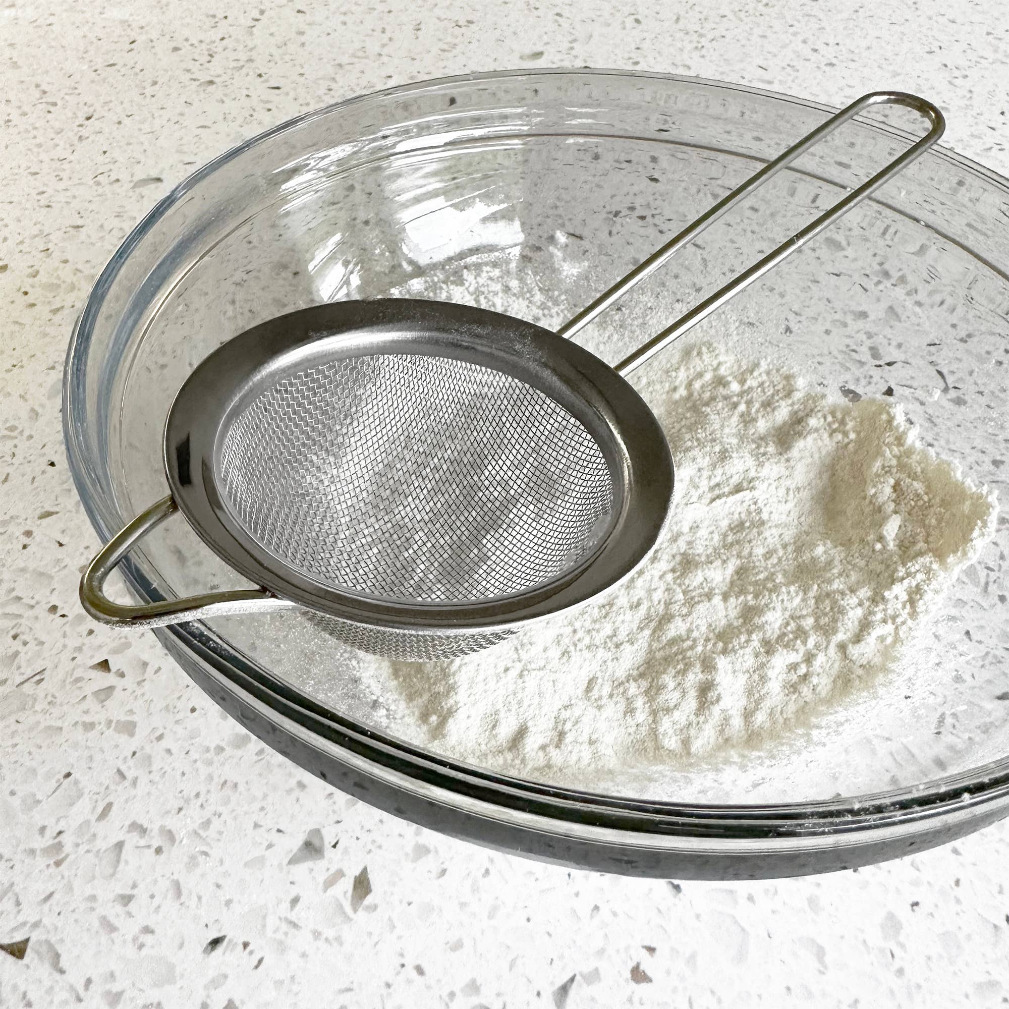 A flour sifter over a glass bowl of flower on a kitchen counter.