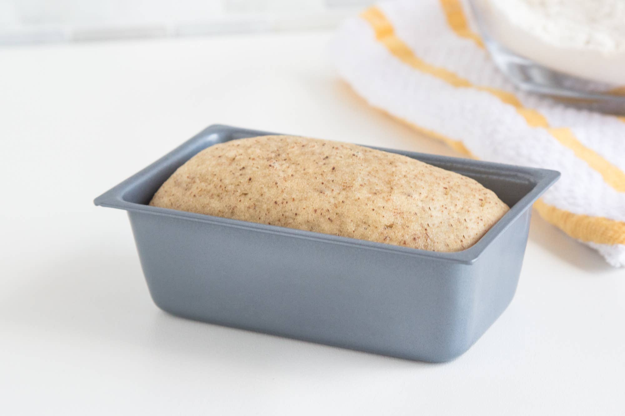 Unbaked bread dough in a loaf pan on a white surface with a yellow and white striped cloth in the background.