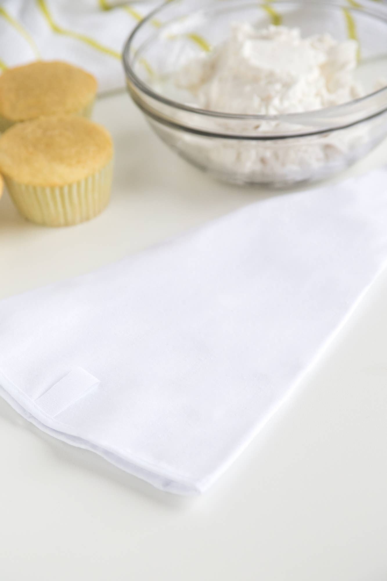 A white reusable icing bag next to a bowl of icing and two cupcakes on a white surface