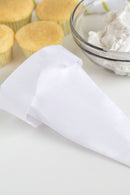 A white reusable icing bag next to a bowl of icing and two cupcakes on a white surface