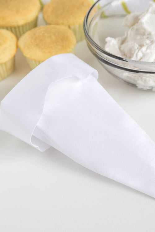 A white reusable icing bag next to a bowl of icing and two cupcakes on a white surface