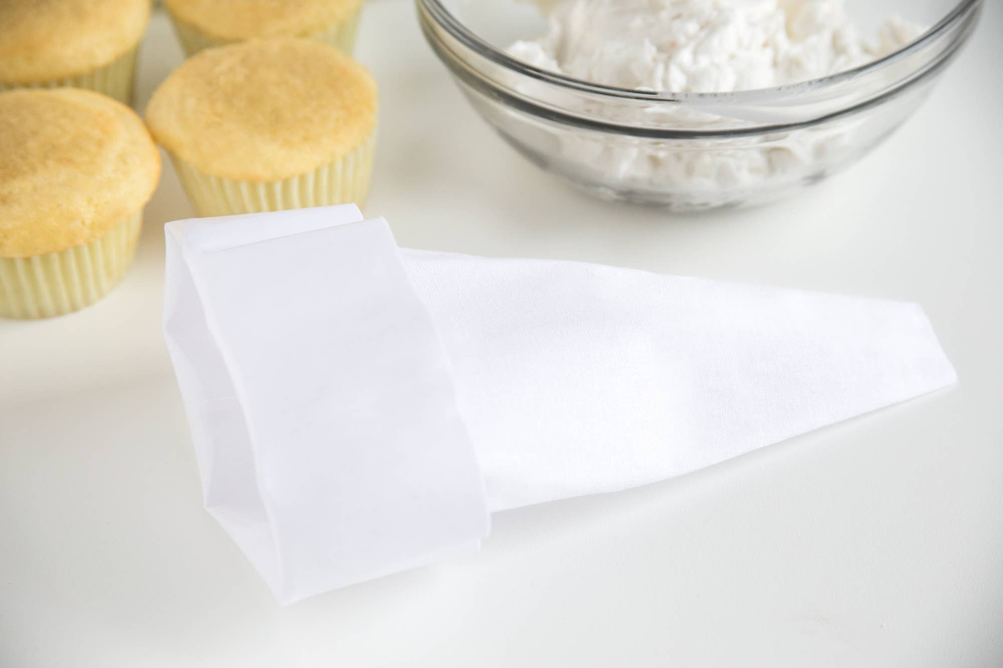 A white reusable icing bag next to a bowl of icing and two cupcakes on a white surface