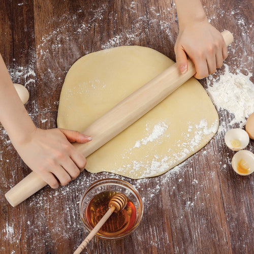 Person rolling out dough on a wooden surface with flour and a bowl of honey.