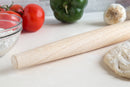 Wooden rolling pin on a white surface with tomatoes, bell pepper, and garlic in the background.