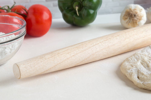Wooden rolling pin on a white surface with tomatoes, bell pepper, and garlic in the background.