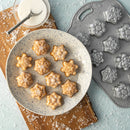 A plate of mini cakes in the shape of snowflakes next to a cake pan with snowflake molds and a bowl of sugar with a spoon on a surface dusted with fake snow.