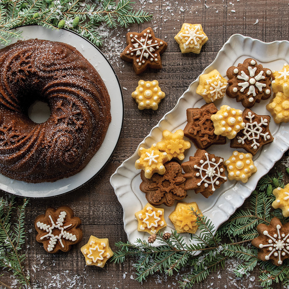 A plate of small cakes shaped like snowflakes and a snowflake themed bundt cake on a dark surface with evergreen branches.