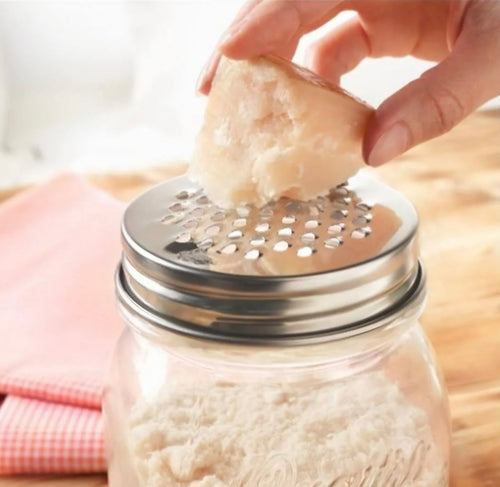A stainless steel grater lid being used on a glass jar, with cheese being grated.