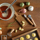 Wooden table with cooking ingredients and utensils including a bowl of sauce, vegetables, and pasta tools.