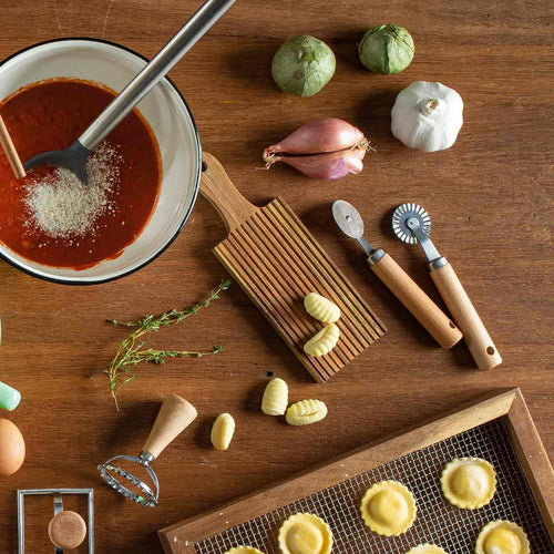 Wooden table with cooking ingredients and utensils including a bowl of sauce, vegetables, and pasta tools.