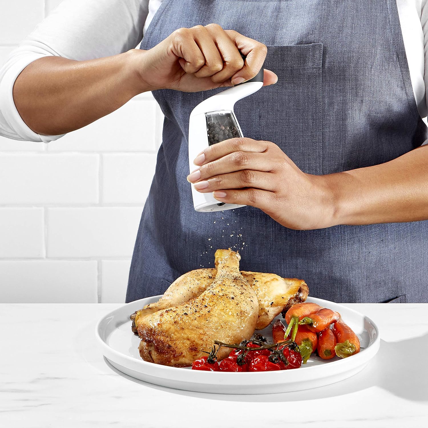 A person using a pepper grinder to put pepper on a meal of chicken, tomatoes, and carrots on a white plate set on a white countertop.
