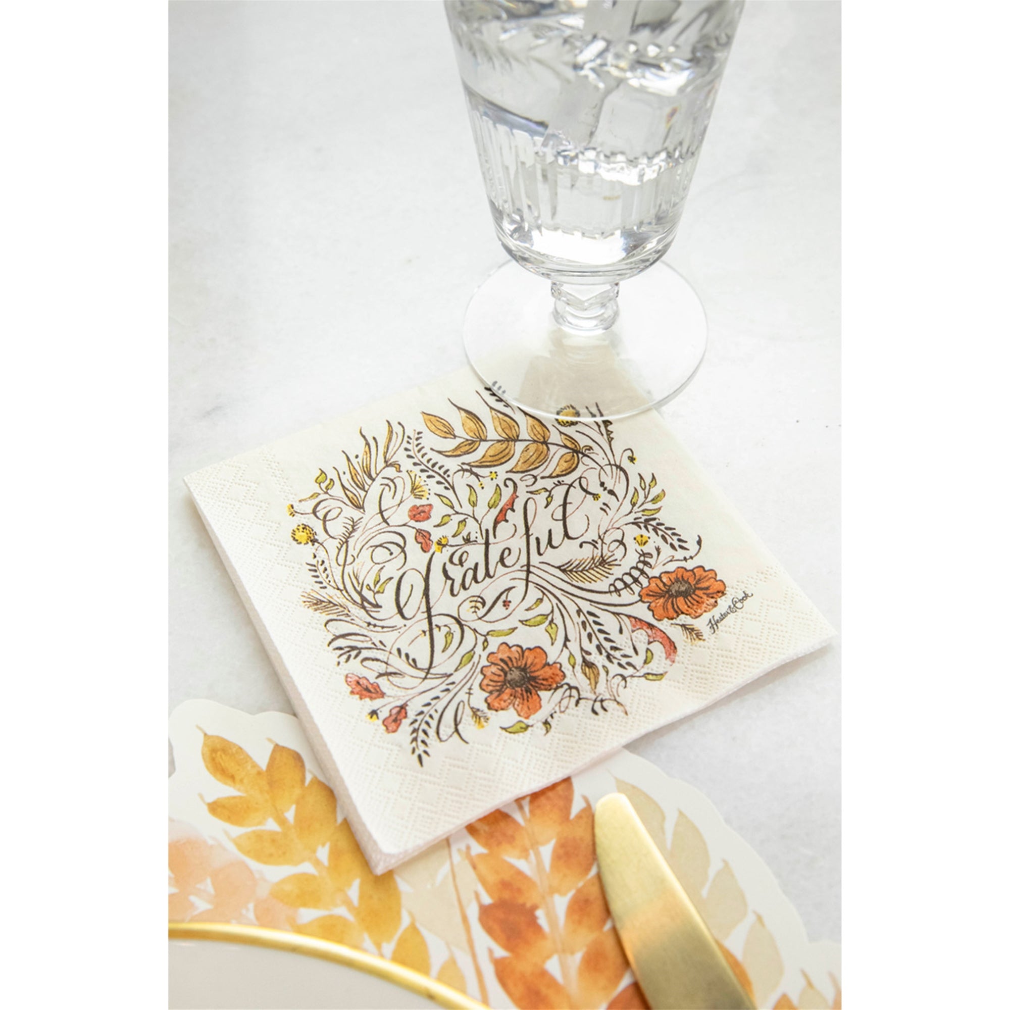 A napkin with autumn artwork and the word 'grateful' next to a paper placemat and a drinking glass on a white surface.