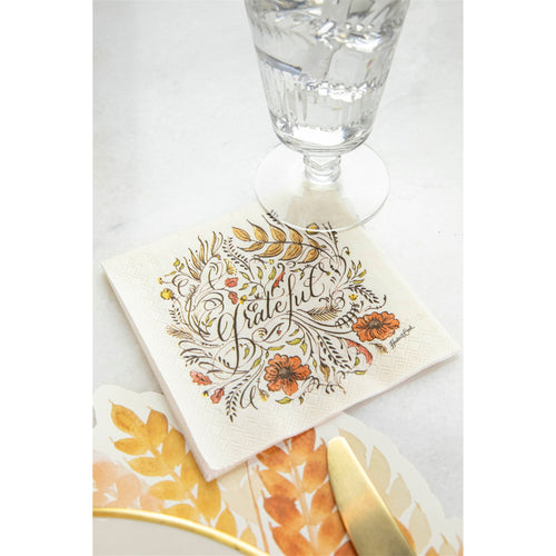 A napkin with autumn artwork and the word 'grateful' next to a paper placemat and a drinking glass on a white surface.