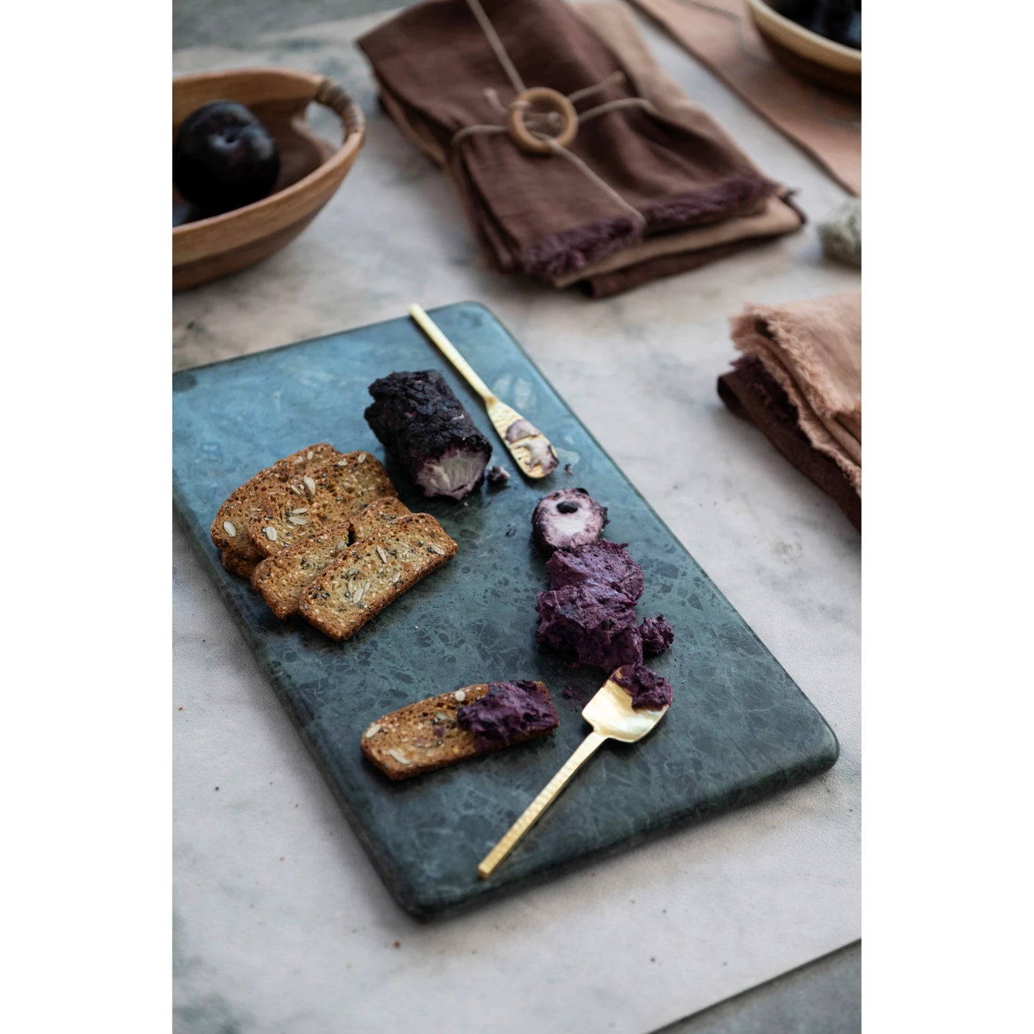 A green marble cutting and serving board on a table with slices of brown bread and a spread with gold utensils.