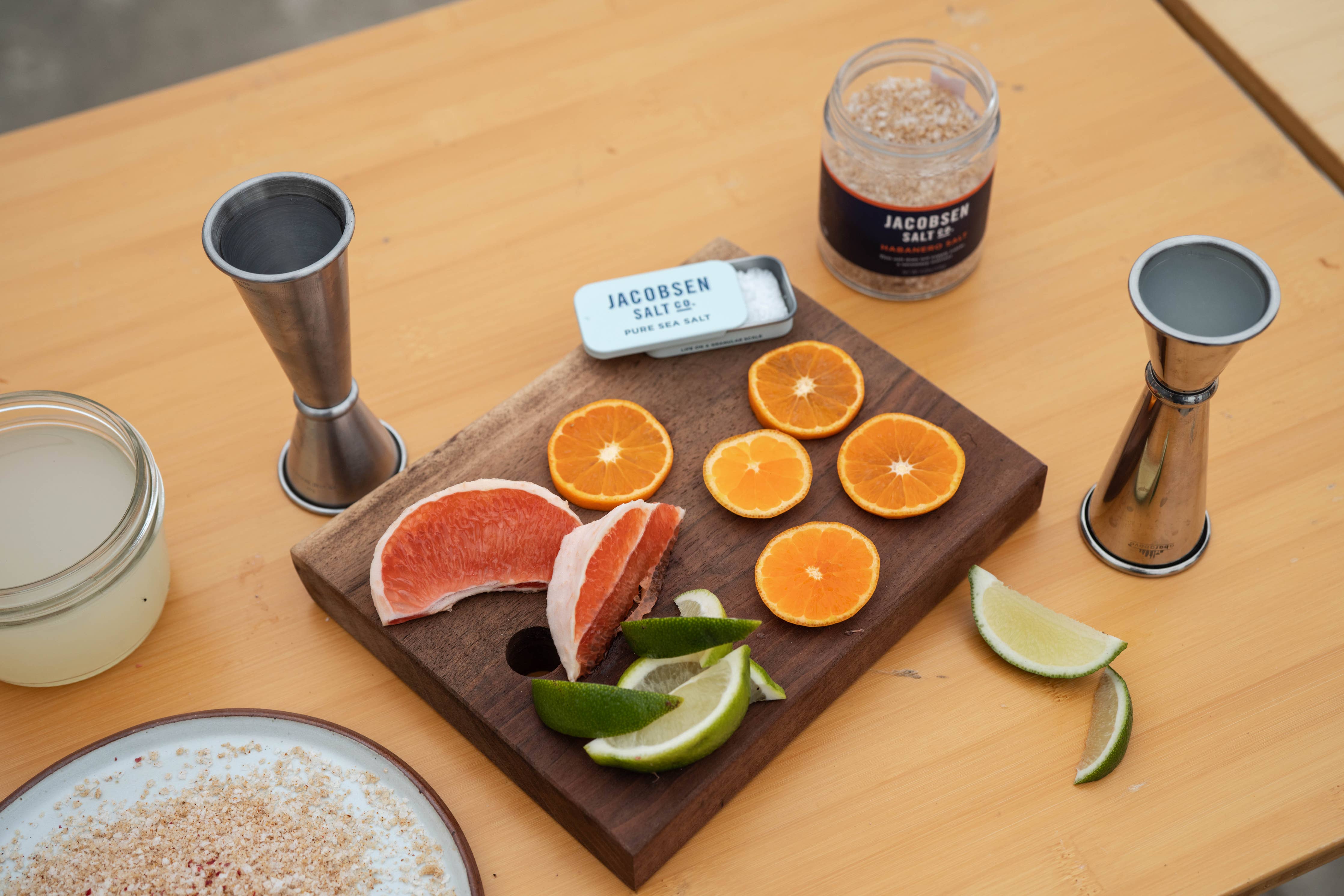 A jar of sea salt with sliced fruit and bar accessories on wooden surfaces.