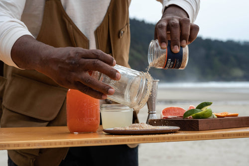 A person using habanero infused sea salt on a glass with liquids and fruit wedges on a wooden surface in an outdoor setting.
