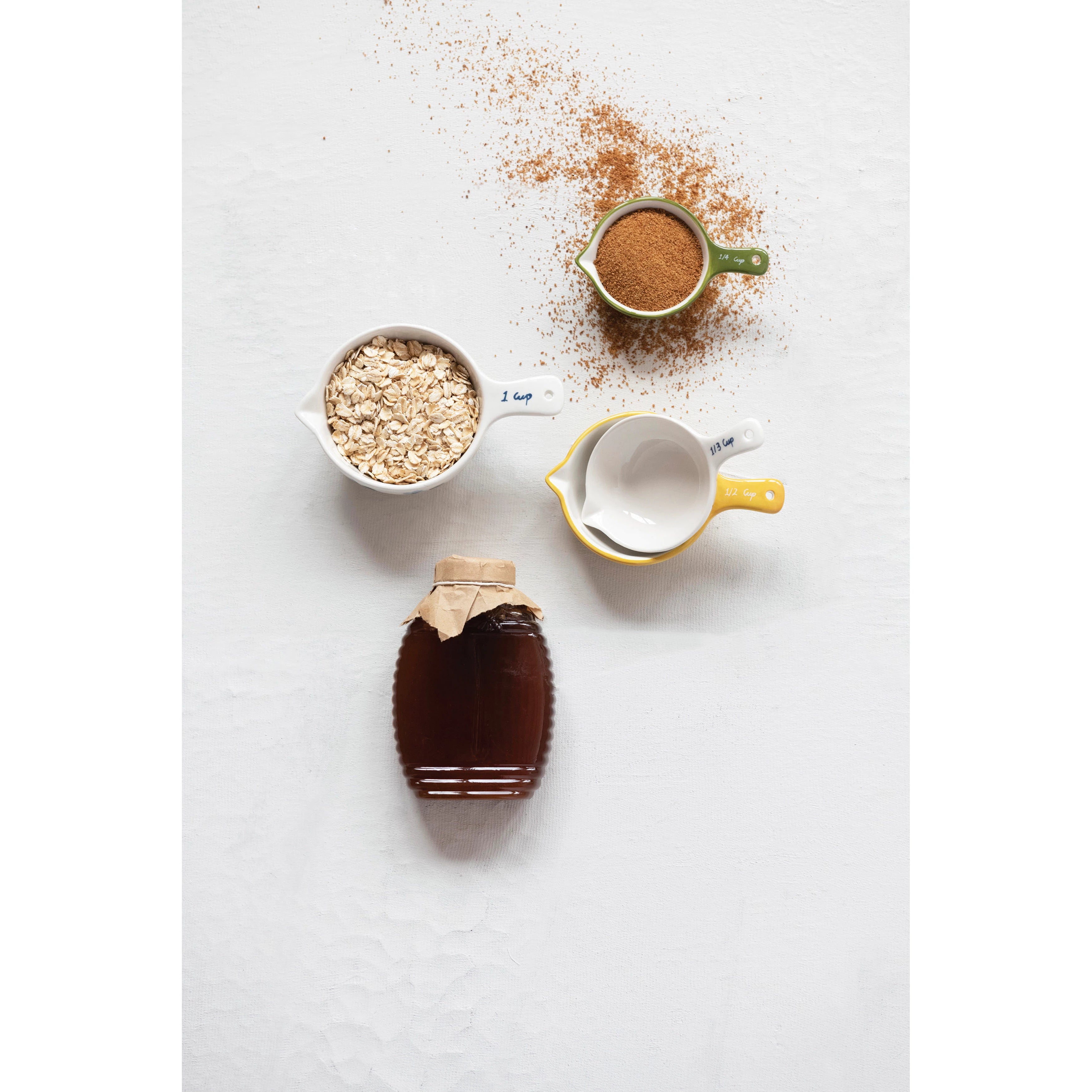 A set of measuring cups with ingredients in two of them next to a container of honey on a neutral surface.