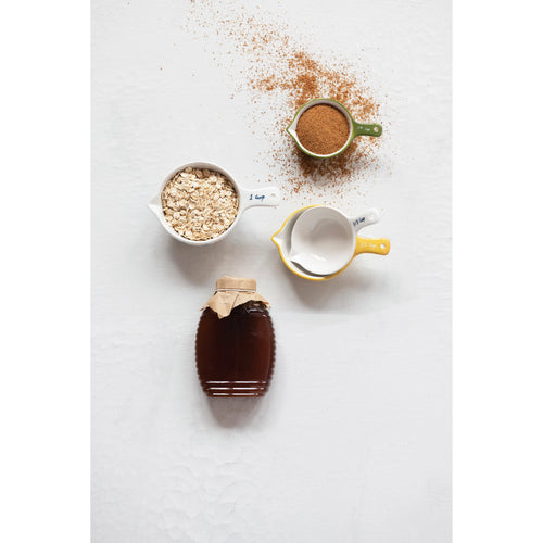 A set of measuring cups with ingredients in two of them next to a container of honey on a neutral surface.