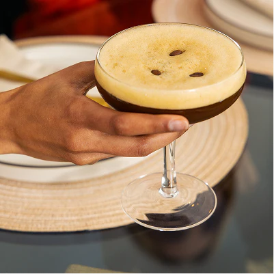 Hand holding a cocktail with an espresso martini on a reflective surface.
