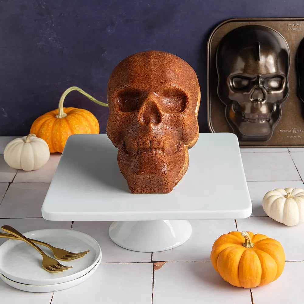 Skull-shaped baked good on a white stand with pumpkins and a baking tray in the background.
