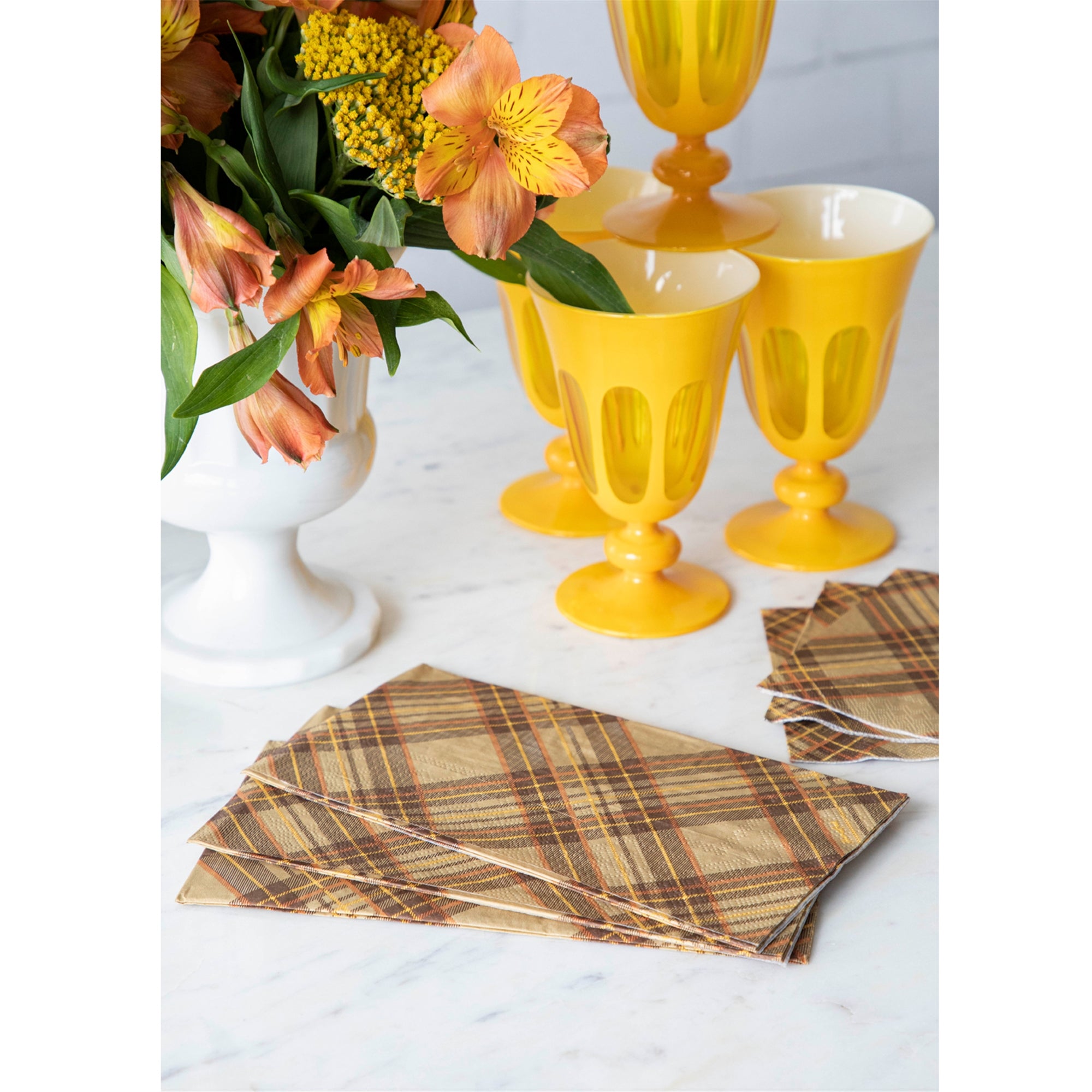 Brown, orange, and yellow plaid napkins on a marble surface with yellow glasses and a white vase with flowers.