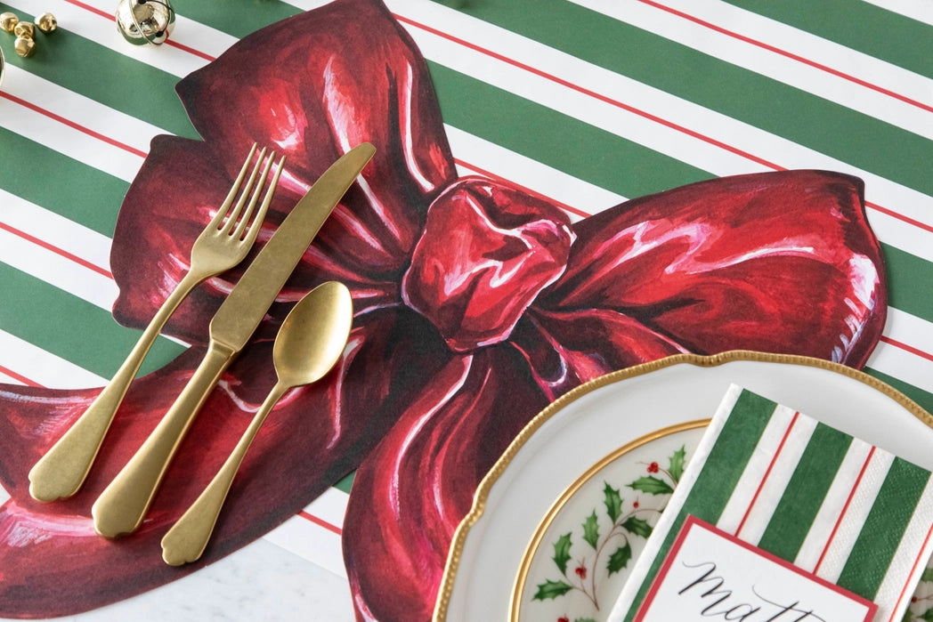 A large red bow shaped placemat at a table setting with gold flatware and a striped table runner.