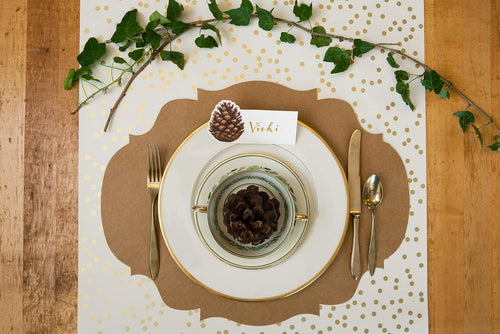 A gold dotted table runner on a wooden surface featuring a French frame placemat with plates and flatware with a sprig of ivy for decoration.