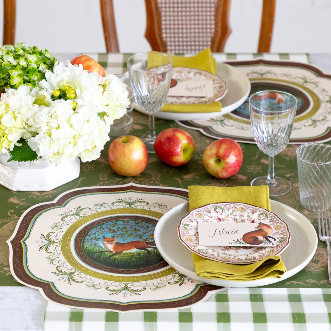 Decorative table setting with floral arrangement, apples, and patterned plates on a checkered tablecloth.