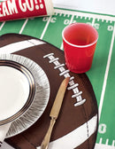 A die-cut football-shaped placemat under a red cup and a brown plate with a football design, featuring a realistic look with marked yard lines and a mock-leather texture.