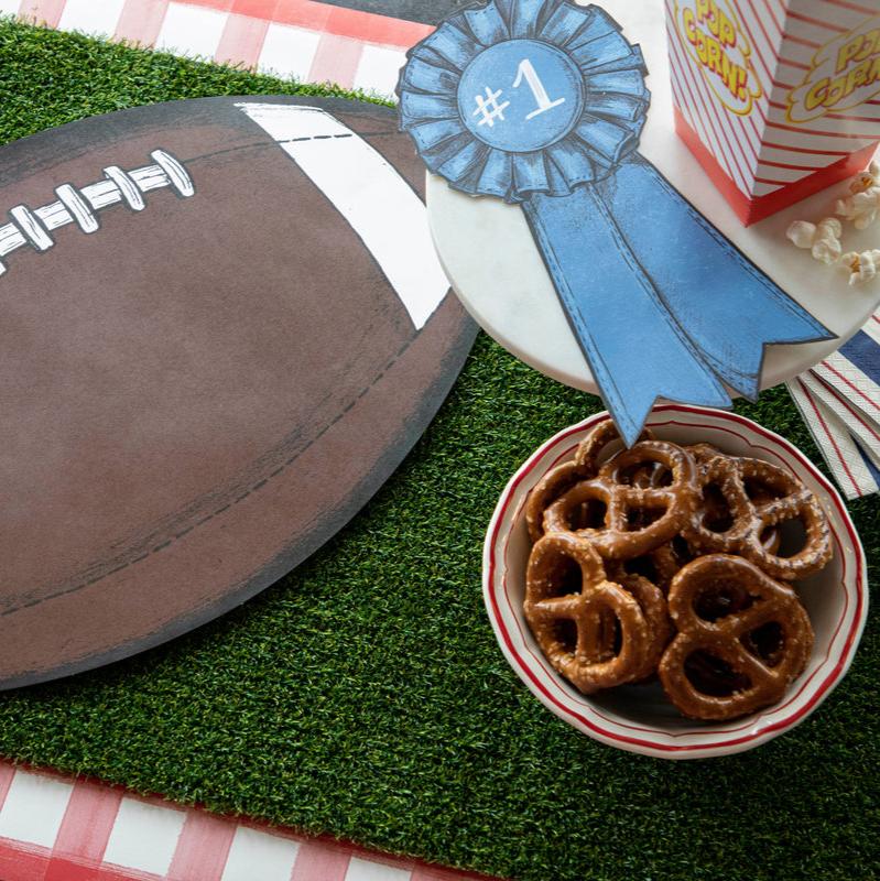 A football shaped placemat on an astroturf-like surface with popcorn, a blue ribbon tag, and a bowl of pretzels.