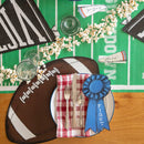 A football shaped placemat on a table with other football themed decorations, popcorn, glasses, and a place setting.