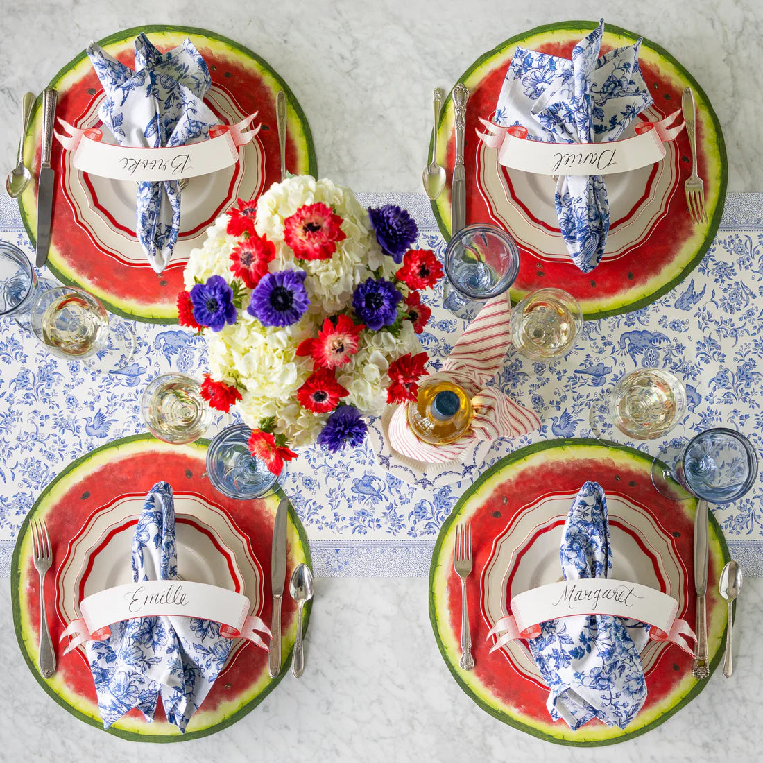 Four place settings on a table with a blue and white table runner, paper watermelon placemats, name cards, drinkware, flatware, plates, blue and white napkins, and floral centerpiece.