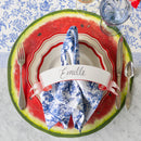 A place setting featuring a paper watermelon slice placemat, plates, flatware, a name card, and blue and white napkin on a blue and white surface.