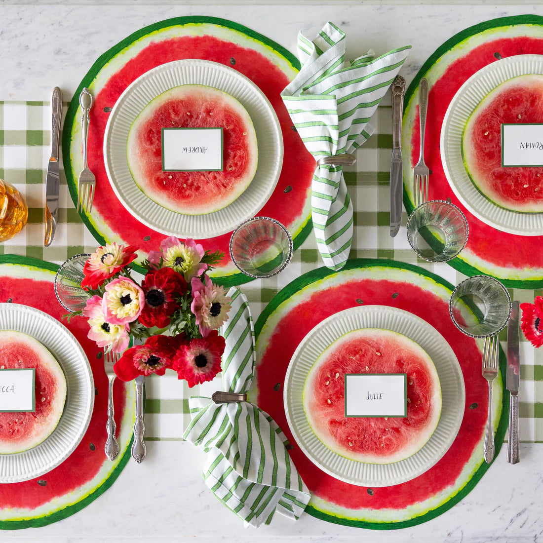 A tablescape featuring place settings with name cards, silver flatware, paper watermelon placemats, green plaid table runner, water glasses, and flowers.