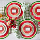 A tablescape featuring place settings with name cards, silver flatware, paper watermelon placemats, green plaid table runner, water glasses, and flowers.