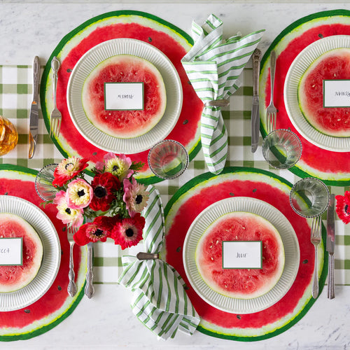 A tablescape featuring place settings with name cards, silver flatware, paper watermelon placemats, green plaid table runner, water glasses, and flowers.