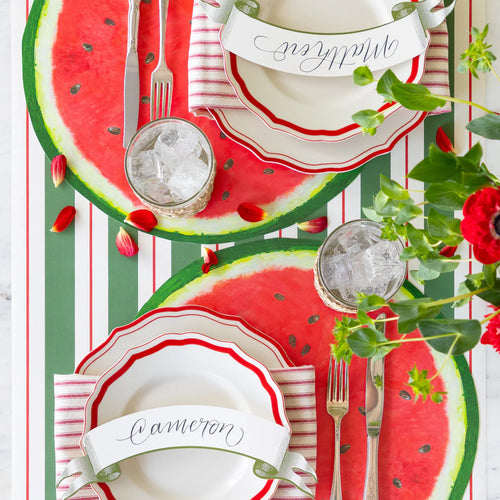 Two place settings featuring name cards, silver flatware, a paper watermelon placemat, and striped table runner.