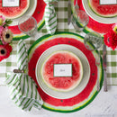 A place setting featuring a name card, silver flatware, green and white striped napkin, a paper watermelon placemat, and green plaid table runner.