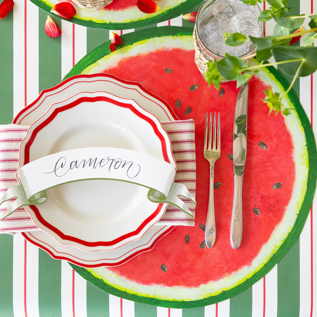 A place setting featuring a name card, silver flatware, a paper watermelon placemat, and striped table runner.