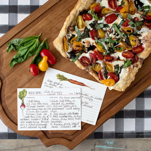 A square pizza topped with cherry tomatoes, basil, and drizzle of oil, served on a wooden board with a handwritten recipe card and fresh tomatoes and basil nearby.