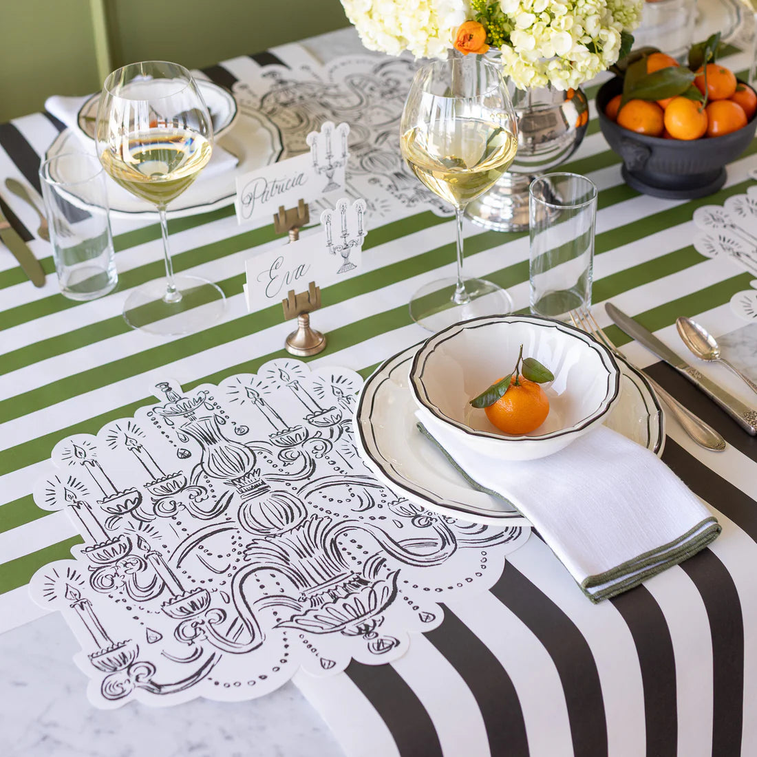 A table setting with chandelier art paper placemats, green striped table runner, black and white striped table runner, glasses of wine and oranges in a bowl.